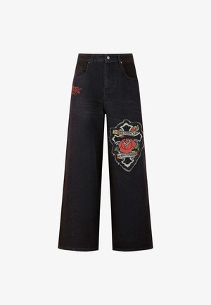 Černé široké džíny s vyšitým červeným růžovým nášivkem a textem "NA PAMĚŤ, KDY JSEM SE ZAJÍMAL." Obsahují klasický design s pěti kapsami a poutky na opasek.