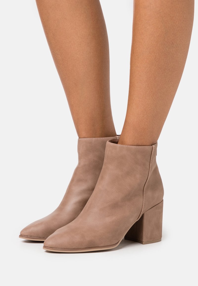 Bottines en daim beige avec talons blocs portées sur jambes nues sur un fond blanc uni.