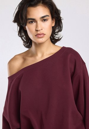 Jong persoon met kort donker haar, die een bordeauxrode, off-shoulder trui draagt en neutraal recht in de camera kijkt.