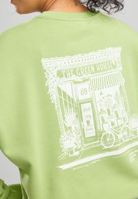 Light green sweatshirt featuring a white illustration of a storefront with flowers, bicycles, and a sign reading "Open."