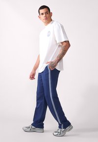 Hvit bomulls T-skjorte med rund grafisk logo, kombinert med marineblå joggebukser med hvite sidestriper og grå joggesko.