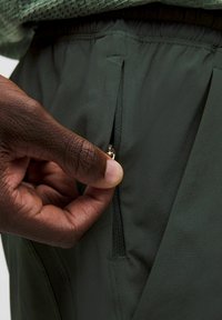 Mörkgröna sportbyxor med en dragkedjeficka, slät textur, elastisk midja och en hand som sträcker sig för att öppna dragkedjan.