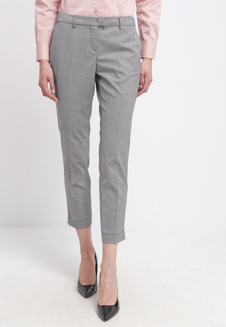Femme portant un pantalon gris moulant à motifs aux chevilles, une chemise rose pâle boutonnée et des talons hauts noirs pointus, debout devant un fond blanc.