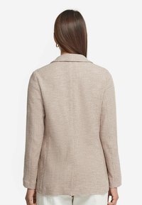 Beige texturerad blazer med slagkrage, långa ärmar och en figurnära design. Har ett diskret mönster och inga synliga knäppningar.