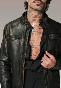 Veste en cuir noire avec deux poches zippées, finition texturée et design ajusté. Portée par-dessus une chemise noire, mettant en valeur des tatouages complexes sur la peau.