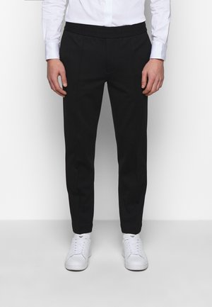 Mężczyzna ubrany w czarne, dopasowane spodnie, białą koszulę i białe trampki, stojący na tle jasnego, jednolitego tła.