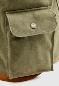 Πράσινη τσάντα καμβά με μια τσέπη με καπάκι, κλειστή με μεταλλικό κουμπί, διαθέτοντας καφέ δερμάτινη βάση και ανθεκτικό υφασμάτινο ανάγλυφο υλικό.