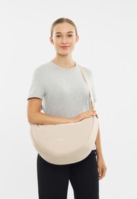 Beige színű, kerek formájú, puha anyagból készült keresztpántos táska, diszkrét márkájú logóval és állítható pánttal.