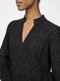 Black embroidered dress with a V-neckline, featuring intricate cut-out patterns and a ruffled collar. Soft, textured fabric, long sleeves.