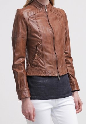 Femme portant une veste en cuir marron cintrée avec une fermeture éclair à l’avant et des poches, associée à un haut sombre et un pantalon blanc.
