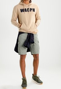 Felpa beige con testo navy "WACPH", abbinata a pantaloni cargo verde chiaro. Le sneakers sono verde scuro con suole bianche e una camicia a quadri è legata in vita.