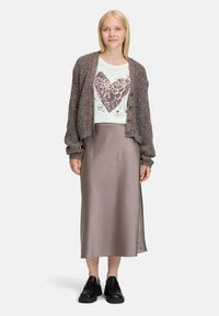 Bruine getextureerde cardigan over een wit T-shirt met een luipaardhartprint, gecombineerd met een satijnen beige rok en zwarte stevige schoenen.