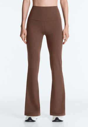 Hnedé slim-fit nohavice s vysokým pásom a miernym rozšírením, nosené s bielymi teniskami, zobrazené spre predok na stojacej osobe.