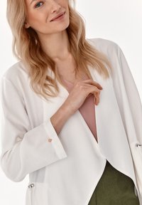 Biely otvorený blazer s dlhými rukávmi, ktorý má jemné ružovo-zlaté akcenty na manžetách a mäkký ružový top pod ním. Viditeľné sú aj zelené nohavice.
