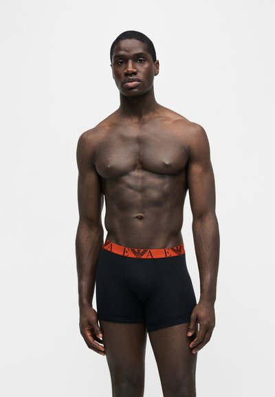Μυώδης άνδρας όρθιος, φοράει μαύρα boxer briefs με κόκκινη μέση με λογότυπα της μάρκας, μπροστά σε απλό ανοιχτό γκρι φόντο.