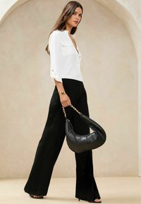 Blusa bianca con colletto e dettagli a bottoni, dotata di due tasche sul petto, abbinata a pantaloni neri a vita alta con gamba wide leg. Viene tenuta in mano una borsa in pelle nera.
