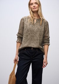 Blonde vrouw draagt een beige blouse met zwarte vlekken en een sleutelgat halslijn, donkere spijkerbroek, en houdt een tan suède handtas vast tegen een effen achtergrond.