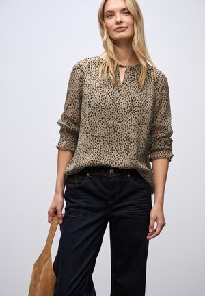 Donna bionda che indossa una blusa beige con macchie nere e scollatura a chiave, jeans scuri, mentre tiene una borsa a spalla in suede color beige su uno sfondo semplice.