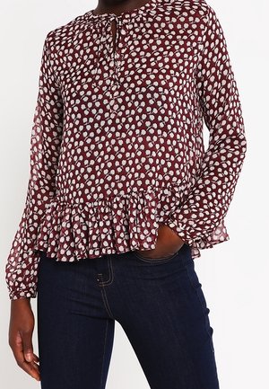Vrouw draagt een bordeauxrode blouse met lange mouwen, wit bloemenpatroon en peplum zoom, gecombineerd met donkerblauwe spijkerbroek.