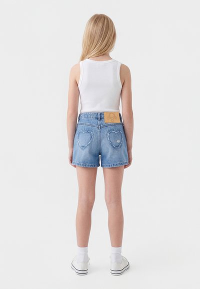 Copil în picioare cu fața spre spate, purtând un maiou alb, pantaloni scurți din denim cu buzunare în formă de inimă pe spate, șosete albe și adidași albi.