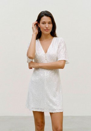 Une femme pose devant un fond clair uni, vêtue d’une robe blanche à sequins, à manches courtes et à col en V.