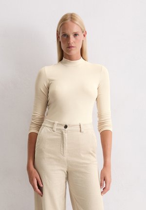 Blonde vrouw draagt een strakke beige coltrui en hoge crèmekleurige corduroy broek, staand tegen een eenvoudige witte muur.