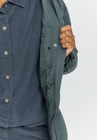 Green padded jacket with a smooth finish, zippered closure, and snap buttons. Worn over a textured navy corduroy shirt with visible buttons.