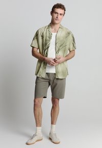 Korte mouwen, lichtgroen shirt met een botanische print; gecombineerd met grijze shorts; model draagt een witte tanktop en beige sneakers.
