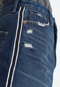Tmavě modré džíny z denimu s bílými a černými bočními proužky, s opotřebovanými detaily a viditelným šitím. Součástí jsou měděné doplňky.
