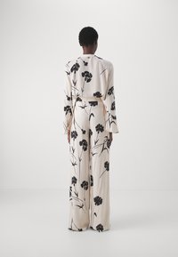Crème-kleurige jumpsuit met zwart bloemenpatroon; heeft brede, lange mouwen en een relaxte pasvorm. Een gladde stof met een zachte textuur.