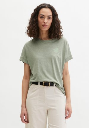 Frau mit lockigem Haar, die ein hellgrünes T-Shirt trägt, das in beige Hosen mit schwarzem Gürtel gesteckt ist, stehend vor einem einfarbigen Hintergrund.