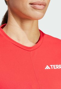 Röd atletisk tröja med rund hals, gjord av mjukt material. Har en vit logotyp och kontrasterande sömdetaljer längs halsringningen.