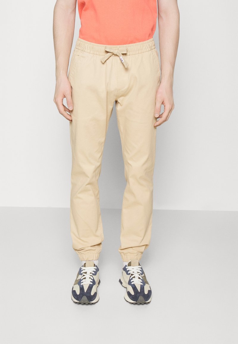 Homme portant un pantalon de jogging beige à cordon, un haut orange et des baskets beige et marine, debout devant un fond uni clair.