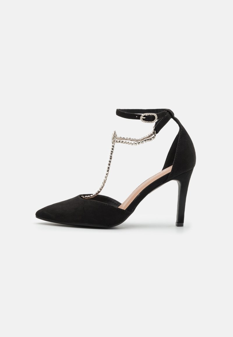 Chaussure noire à talon haut avec bout pointu, bride de cheville à boucle et chaîne décorative en strass reliant le bout du pied à la bride de cheville.