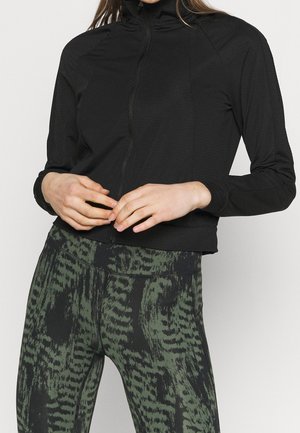 Person trägt schwarze Jacke mit Reißverschluss und grüne gemusterte Leggings, steht mit verschränkten Händen auf der Taille vor einfarbigem Hintergrund.