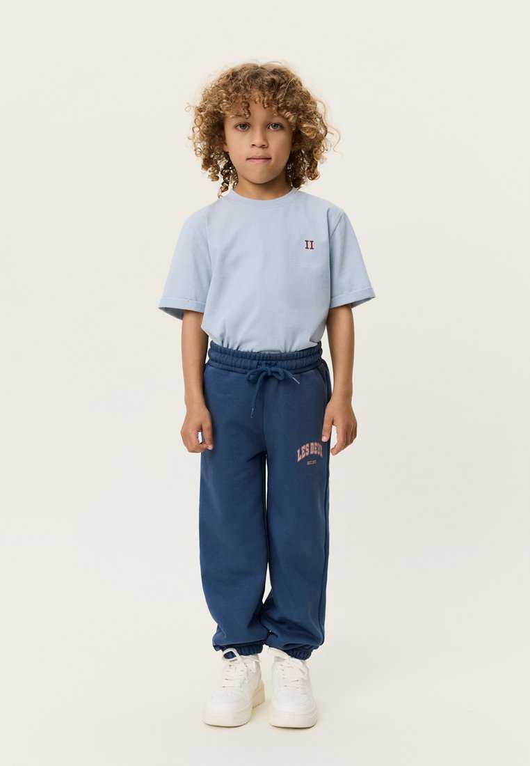 Ανοιχτό μπλε κοντομάνικο πουκάμισο με ένα μικρό κόκκινο γραφικό, συνδυασμένο με σκουρό μπλε φόρμα με ροζ λογότυπο και ελαστικές μανσέτες. Λευκά αθλητικά παπούτσια.