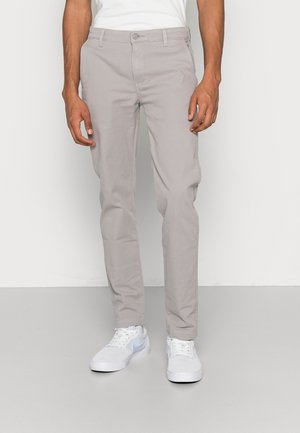 Uomo con pantaloni aderenti grigio chiaro e sneakers bianche, in piedi davanti a uno sfondo chiaro e uniforme.