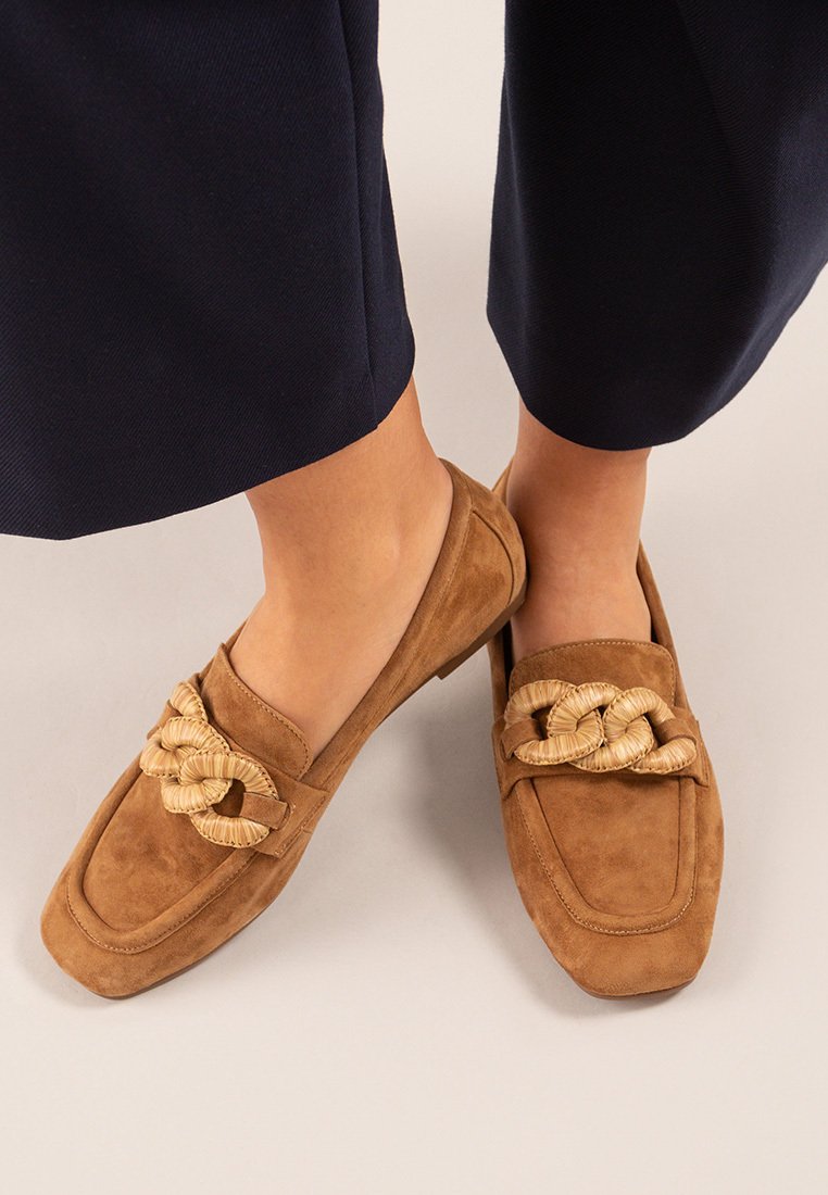 Mocasines de ante en tono marrón claro con un acento de paja trenzada en la parte delantera, presentando un diseño de punta cuadrada y un tacón bajo, combinados con pantalones oscuros.