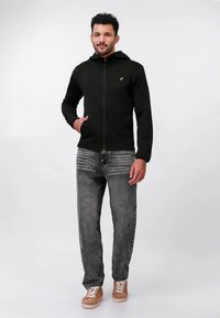 Uomo che indossa una felpa nera con zip, jeans grigi lavati e sneakers marroni, in piedi davanti a uno sfondo bianco uniforme.