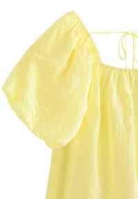 Gul blus med puffärmar, med en texturerad mönster och samlad halsringning. Snörena i ryggen ger detaljer. Ljust, luftigt tyg.