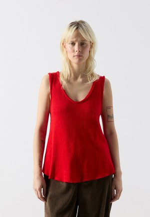 Personne blonde portant un haut rouge sans manches et un pantalon marron, debout devant un fond blanc uni, face à l'avant.