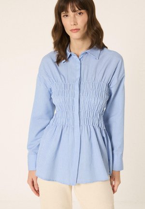 Žena nosi svetlomodrú košeľu s dlhým rukávom a golierom, s bielymi vertikálnymi pásmi a nazbieraným elastickým detailom v páse.