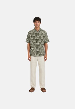 Hombre de pie vestido con una camisa de manga corta verde con estampado, pantalones beige claro y sandalias marrones contra un fondo claro y liso.
