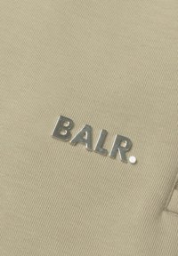 Beige stof met een gestructureerd oppervlak featuring een zilverkleurig metallic 'BALR.' logo, prominent weergegeven op het materiaal.