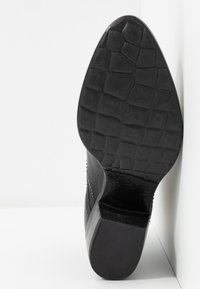 Zapato de tacón negro con una suela texturizada con un patrón de diamantes, adornos de tachuelas a lo largo del lateral y un material superior suave.