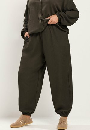 Persoon draagt losse donkergroene textuur broek met elastische boorden en bijpassende top, staat met handen in de zakken en beige casual sneakers.