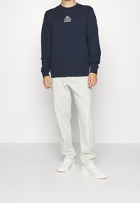 Marinblå sweatshirt med Lacoste-logotyp på bröstet, kombinerad med ljusa randiga byxor och vita sneakers.