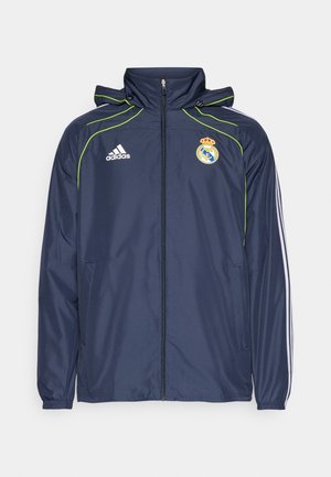Giacca a vento navy con accenti verdi, chiusura zip frontale, cappuccio e tasche laterali. Presenta il logo Adidas e l'emblema del Real Madrid sul petto.