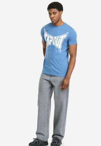 Blaues Baumwoll-T-Shirt mit großem weißem "TAPOUT"-Logo, kombiniert mit grauen Jeans und schwarzen Sneakern. Einfaches Design, lässige Passform.