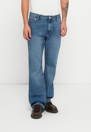 Homme portant un jean bleu clair à coupe décontractée, une chemise blanche et des chaussures en cuir marron, se tenant devant un fond blanc uni.